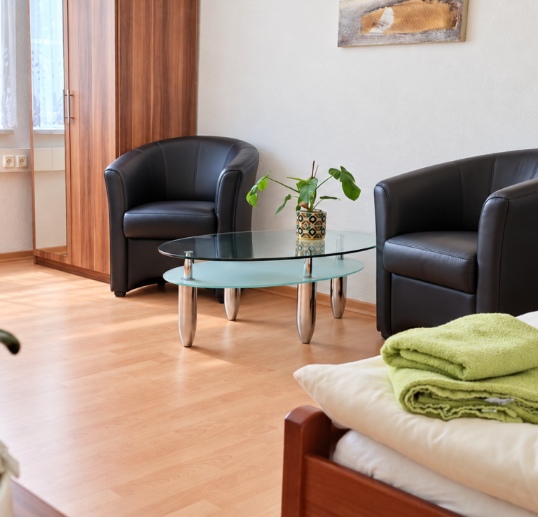Sitzecke Sitzecke mit zwei grauen Sesseln, Glastisch und einer Pflanze neben einem Bett mit grünen Handtüchern.